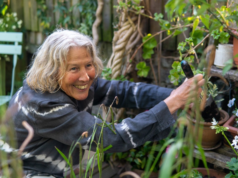 An elderly woman smiling, digging dirt in a pot, surrounded by plants and flowers.